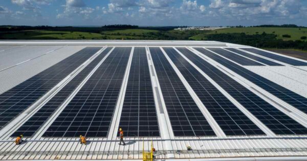 Vista aérea de telhado industrial com extensa instalação de painéis solares e equipe técnica em operação em projeto de energia solar empresarial.